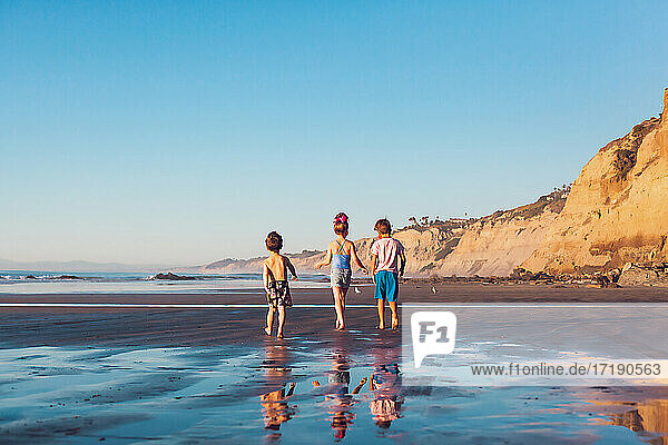 Three little kids walking on the beach at low tide at sunset.