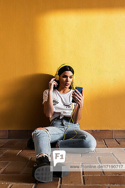 Portrait of Spanish brunette girl sitting on the floor and listening to music with headphones on yellow background wall.