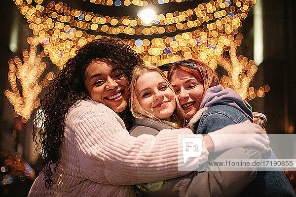 Glückliche Freundinnen  die sich bei der Weihnachtsbeleuchtung in der Stadt umarmen