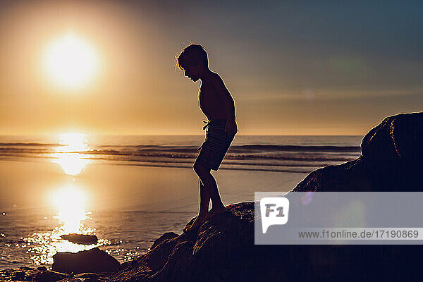 Silhouette eines Jungen  der bei Sonnenuntergang die Felsen am Strand hinunterläuft.