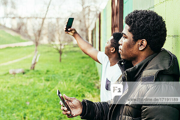 Porträt von zwei schwarzen Jungen  die an einer grünen Wand lehnen. Sie benutzen ihre Handys.