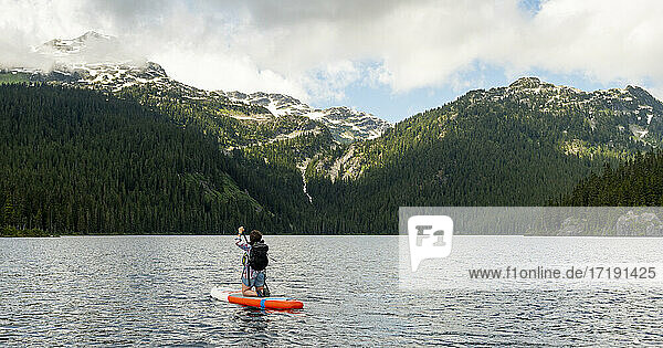 Rückenansicht einer Frau  die auf einem Paddleboard kniet und auf dem kräuselnden Wasser eines Sees in Whistler  Kanada  in Richtung der grünen Berge paddelt