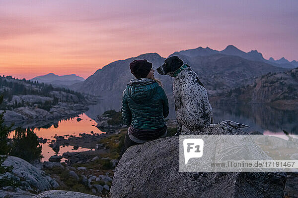 Rückansicht einer jungen Frau  die mit einem Hund auf einem Berg bei Sonnenuntergang auf einem Felsen sitzt