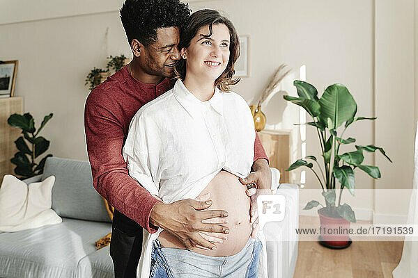 Glücklicher ethnischer Mann  der eine schwangere Frau zu Hause umarmt