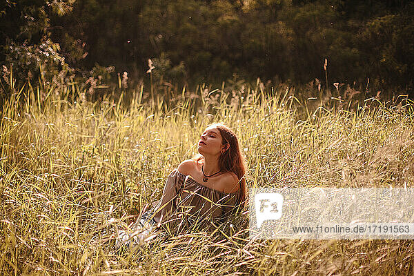 Friedliche junge Frau  die im Sommer auf einem Feld im Gras sitzt