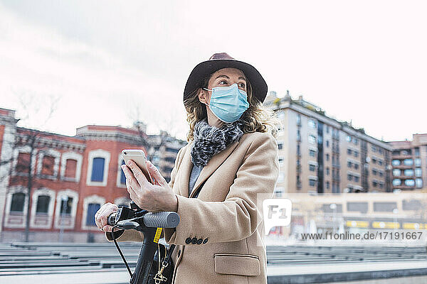 Blond woman with hat and mask  consults her phone and uses her electri
