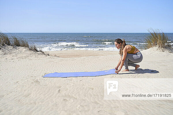Ein hübsches junges Mädchen legt eine Matte an den Strand  um ihre morgendlichen Yogaübungen zu machen.