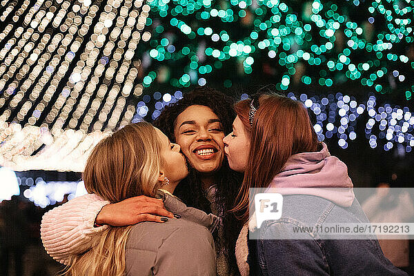 Freundinnen küssen junge Frau auf die Wangen bei Weihnachtsbeleuchtung
