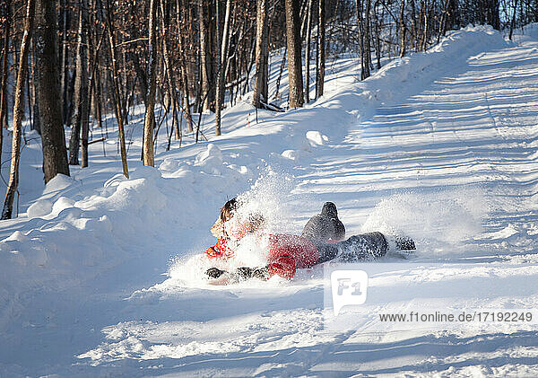 Junges Kind schlittelt einen verschneiten Hügel in einem Waldgebiet an einem sonnigen Tag hinunter.