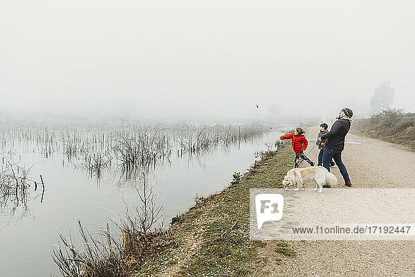 Father and two sons with dog throwing stones into lake on foggy day