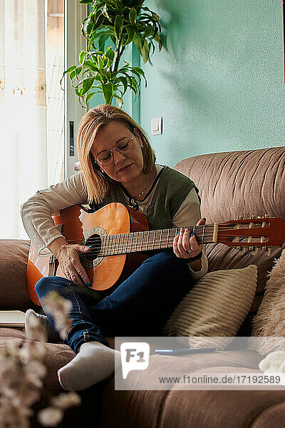 Frau mittleren Alters spielt Gitarre auf dem Sofa im Wohnzimmer zu Hause