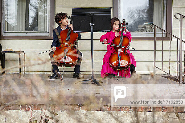 Zwei Kinder sitzen in Kostümen draußen und spielen Cello auf der Veranda