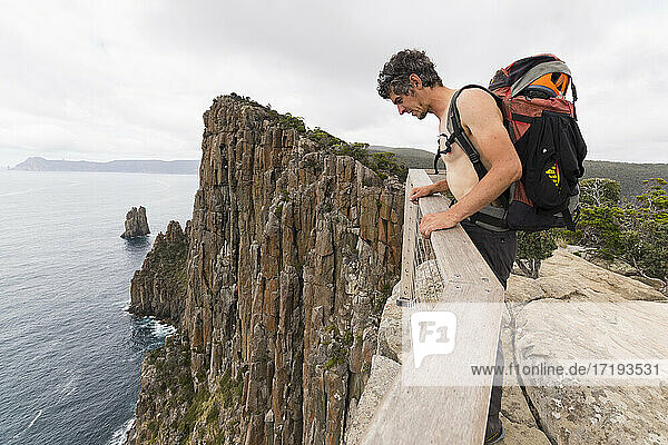 Man carrying a backpack leans over the top of sea cliff in Tasmania.
