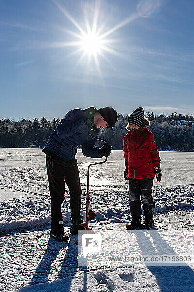 Two boys using auger to make a hole in the ice on a sunny winter day.