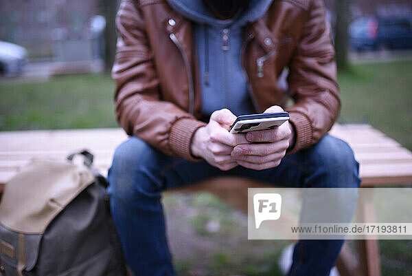 Das Bild konzentriert sich auf die Hände eines Mannes mit seinem Mobiltelefon auf einer Holzbank in einem Park.