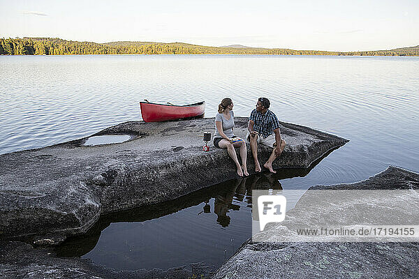 Couple look at view while sitting on rocky lake shore with red canoe