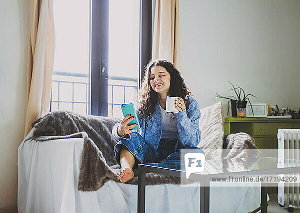 junge Frau beim Frühstück auf dem Sofa mit Kaffee und Handy