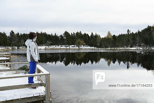 Frau steht auf einer verschneiten Holzbrücke und blickt auf einen zugefrorenen See