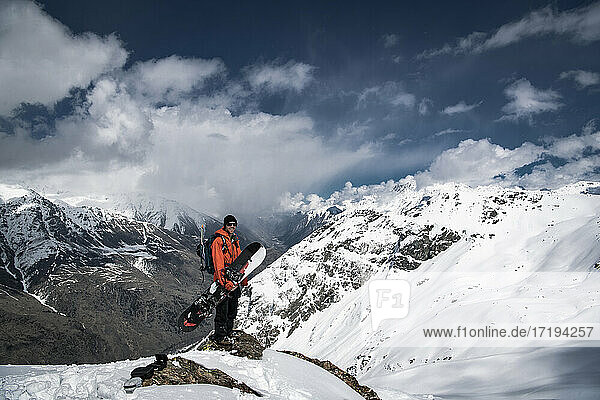 Man with snowboard standing on snowcapped mountain against cloudy sky