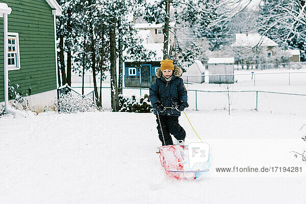 Little happy boy pulling his sled in snowy backyard to go sledding
