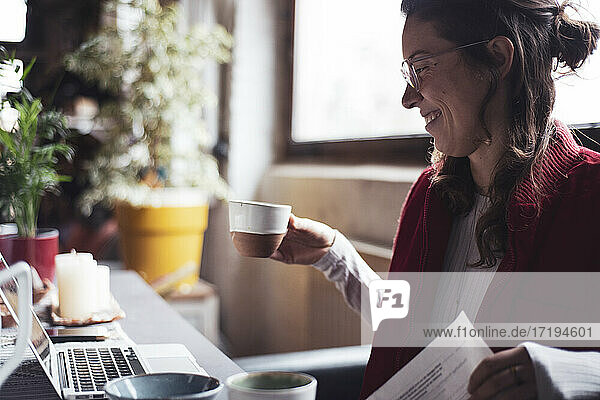 woman smiles with cup of tea while working on laptop in home office