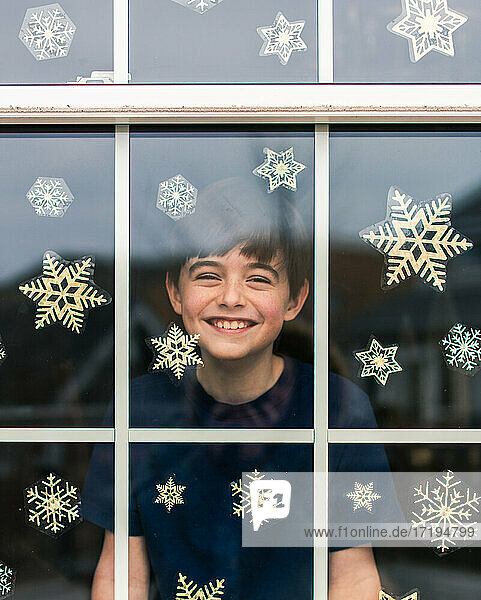 Fröhlicher Junge  der durch ein mit Schneeflocken bedecktes Fenster hinausschaut.