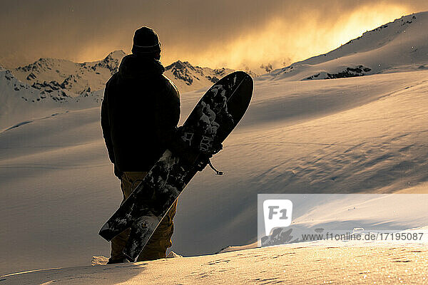 Rückansicht eines Mannes mit Snowboard auf einem schneebedeckten Berg bei Sonnenuntergang