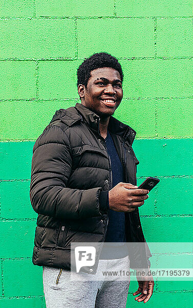 Junger schwarzer afroamerikanischer Junge benutzt sein Mobiltelefon auf grünem Wandhintergrund.