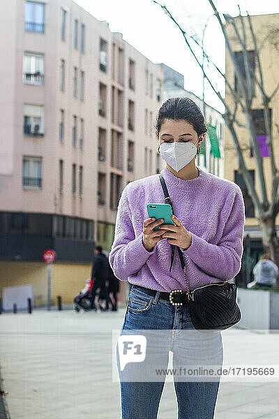 junge Frau mit KN95-Schutzmaske auf der Straße  die das Handy beobachtet