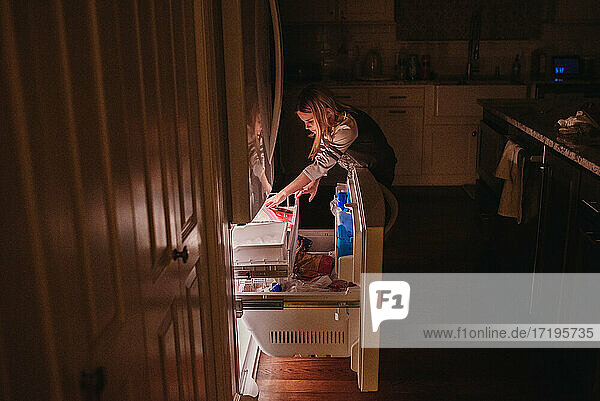 Tween girl reaching into freezer drawer in dark kitchen