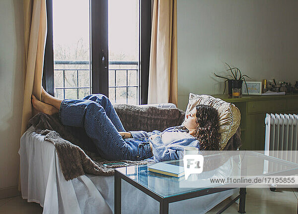 junge Frau mit lockigem Haar  die auf dem Sofa liegt und auf das Fenster schaut
