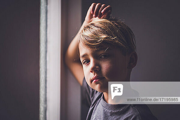 Junge mit haselnussbraunen Augen  der vor einem Fenster steht und in die Kamera schaut.