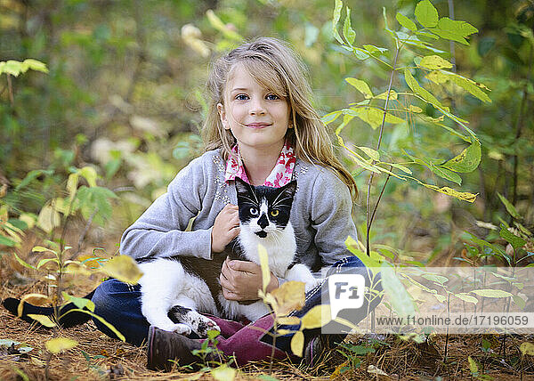 Junges blondes Mädchen sitzt im Wald mit schwarz-weißer Katze auf dem Schoß.