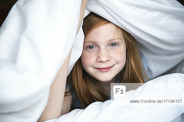 Ein junges rothaariges Mädchen lugt unter einer weißen Bettdecke hervor.