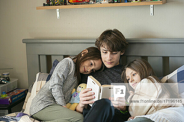 Tween boy reading to his little sisters in bedroom.