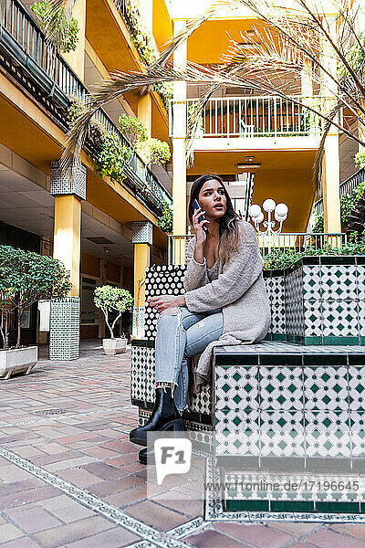 Junge spanische brünette Frau mit Einkaufstüten  die auf einer Bank sitzt und mit ihrem Handy telefoniert.