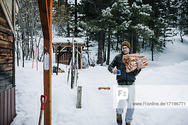 A man wearing flannel carries firewood and a coffee back to cabin