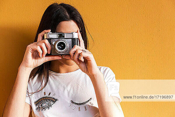 Porträt eines spanischen brünetten Mädchens  das ein Foto mit einer Vintage-Kamera auf einer Wand mit gelbem Hintergrund macht.