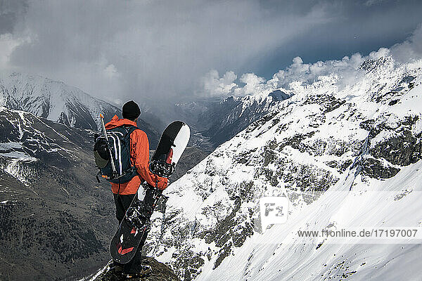Man with snowboard looking at view from snowcapped mountain against cloudy sky