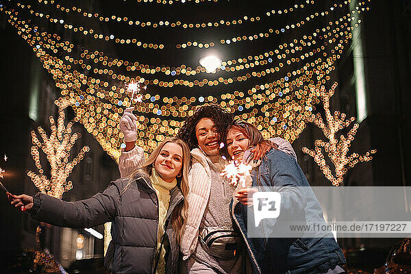 Happy female friends holding sparklers in city during Christmas