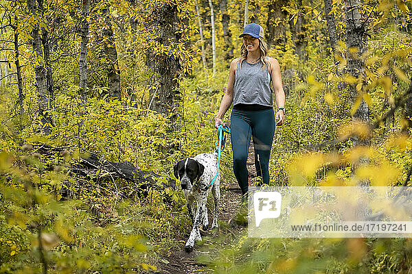 Lächelnde junge Frau beim Spaziergang mit Hund inmitten von Herbstpflanzen im Wald