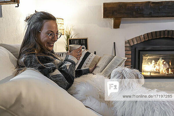 glückliche Frau zu Hause liegend auf Buch lesen und Kaffee trinken mit ihrem Hund