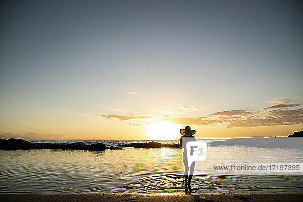 Eine Frau steht in der Untiefe und trägt einen Hut bei Sonnenuntergang
