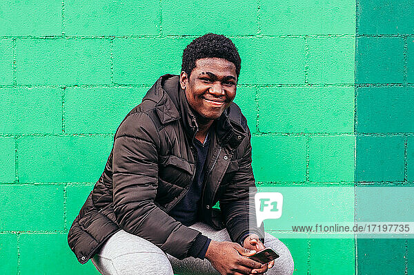 Junger schwarzer afroamerikanischer Junge hockt mit seinem Handy auf grünem Wandhintergrund.