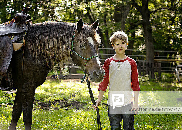 Hübscher blonder Junge  der die Zügel eines Pferdes auf dem Lande hält.
