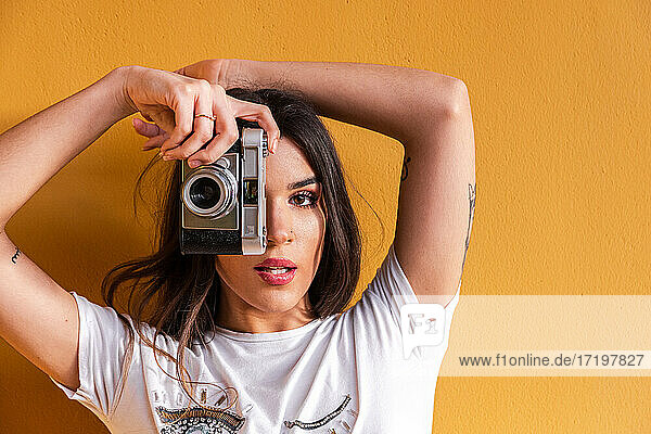 Porträt eines spanischen brünetten Mädchens  das ein Foto mit einer Vintage-Kamera auf einer Wand mit gelbem Hintergrund macht.