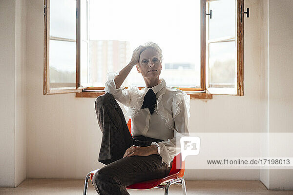 Seriöse Geschäftsfrau mit Hand in den Haaren  die auf einem Stuhl vor dem Fenster in einem Heimbüro sitzt