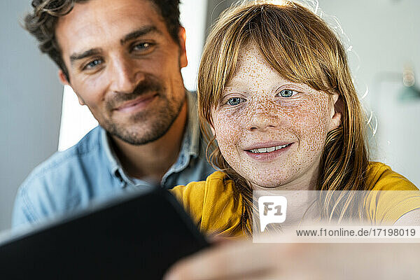 Lächelnder Vater und süße rothaarige Tochter schauen gemeinsam im Wohnzimmer auf ein digitales Tablet