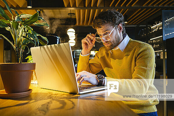 Male owner holding eyeglasses while while working on laptop at cafe