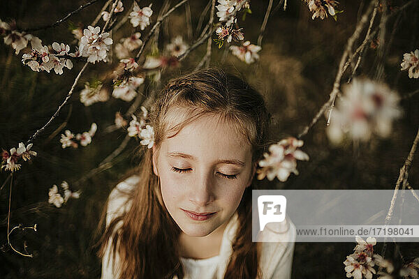 Girl standing with eyes closed under branches of almond tree during springtime